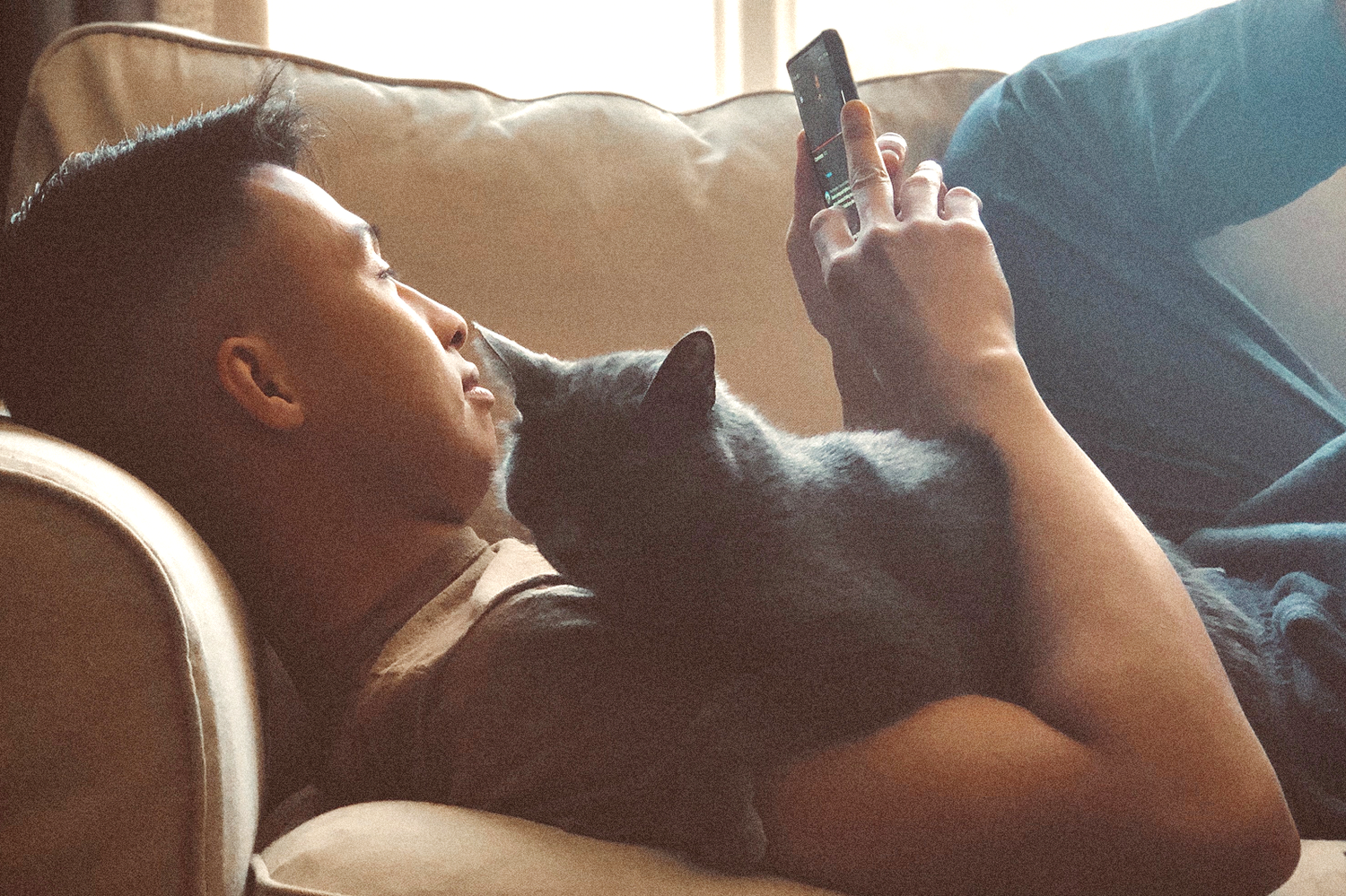 Man laying on a couch using his phone with a black cat sleeping on his chest.