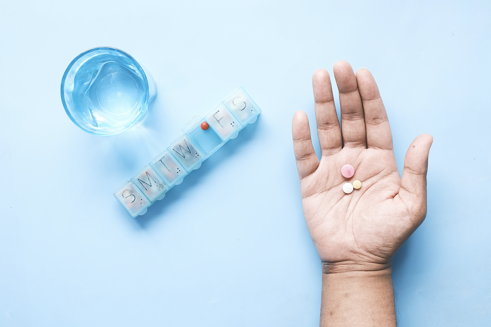 Hand holding pills next to a pill organizer and glass of water.