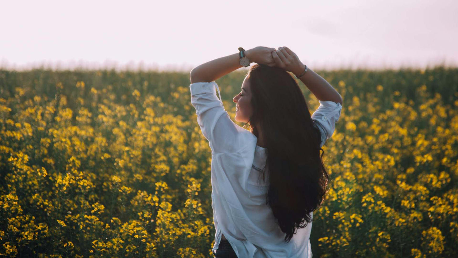 A dark haired woman wearing a white shirt stands before a field of yellow flowers at golden hour with an expression of calm, her arms raised above her head.