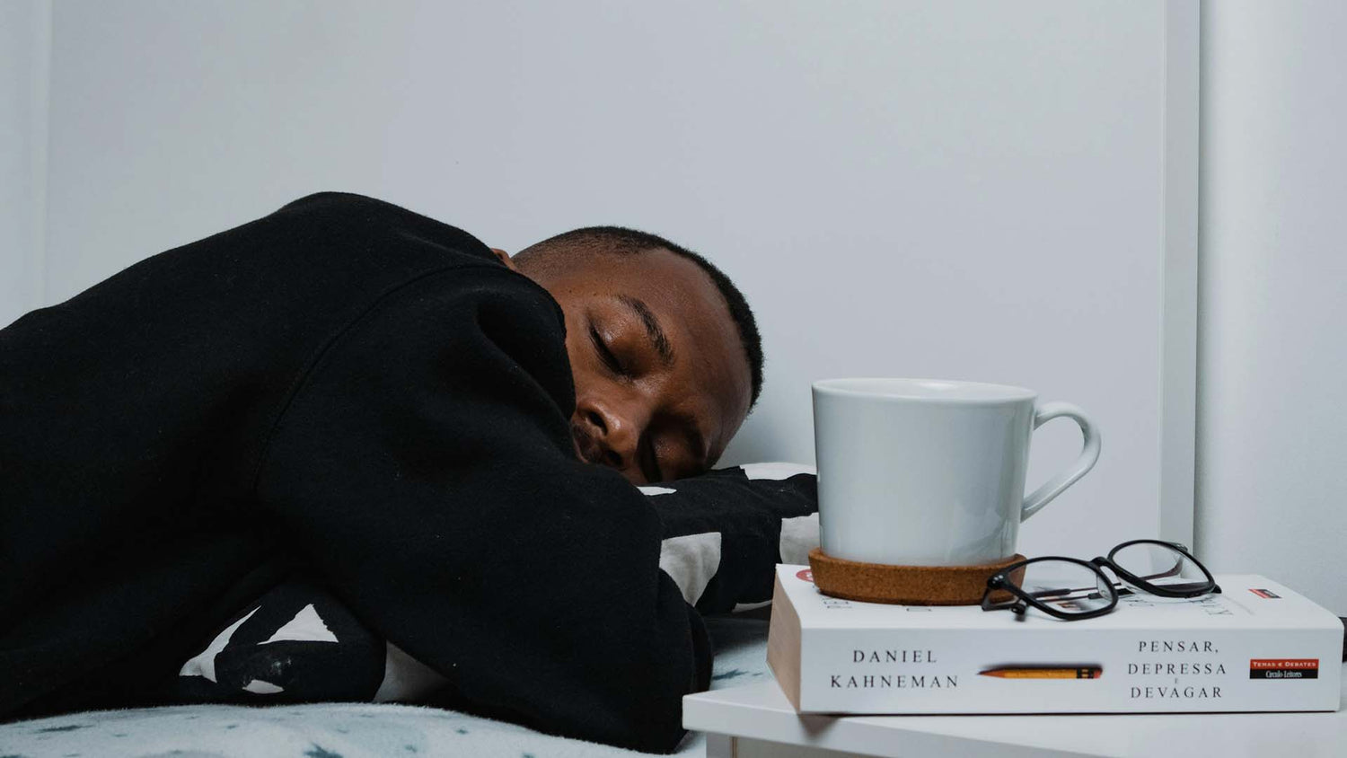 a man sleeping on a bed next to a bedside table with a book and a mug