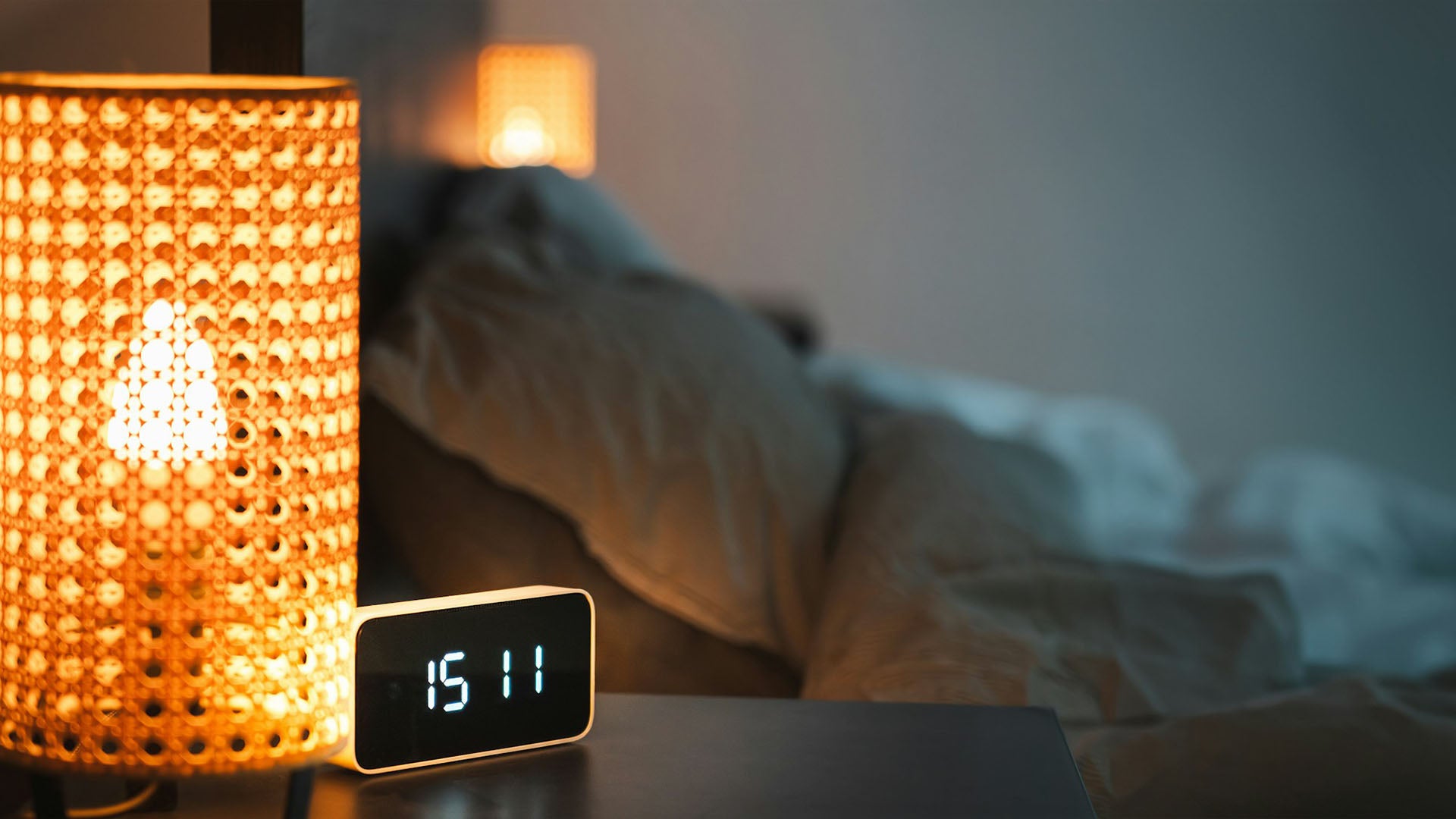 a digital alarm clock sitting on top of a table next to a lamp with a bed in the background