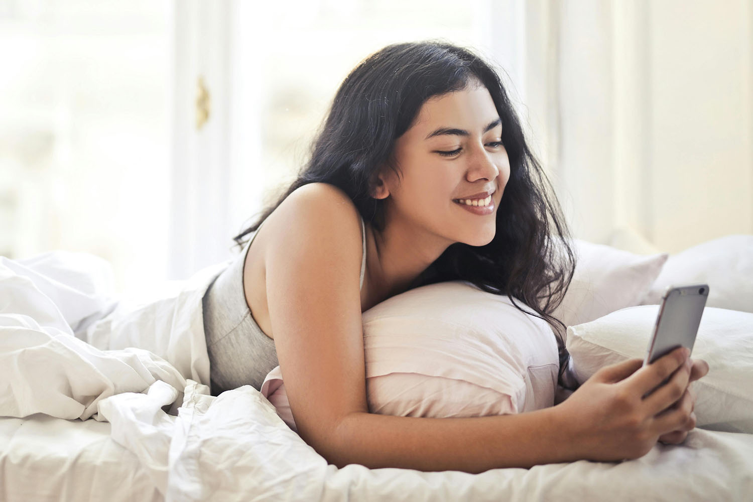 Smiling woman lying on her stomach in bed using a phone